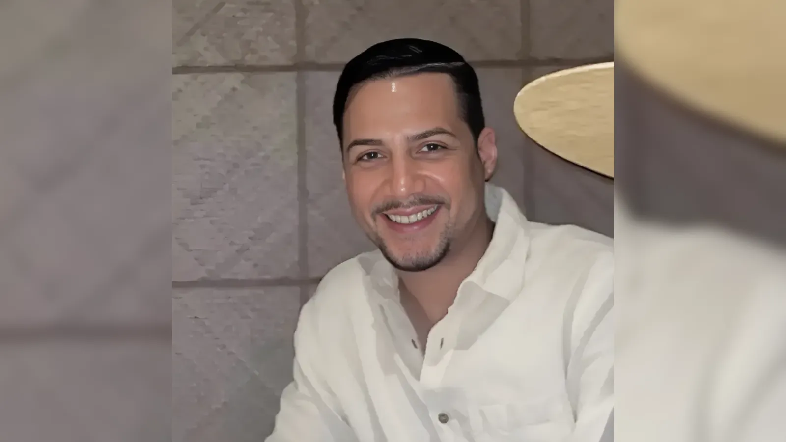 Smiling man with short dark hair in a white button-up shirt, posing indoors in front of a stone wall.