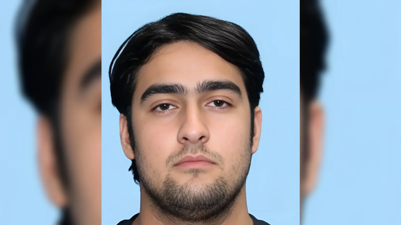 Front-facing head-and-shoulders photo of a young man with dark hair and light stubble against a light blue background, neutral expression.