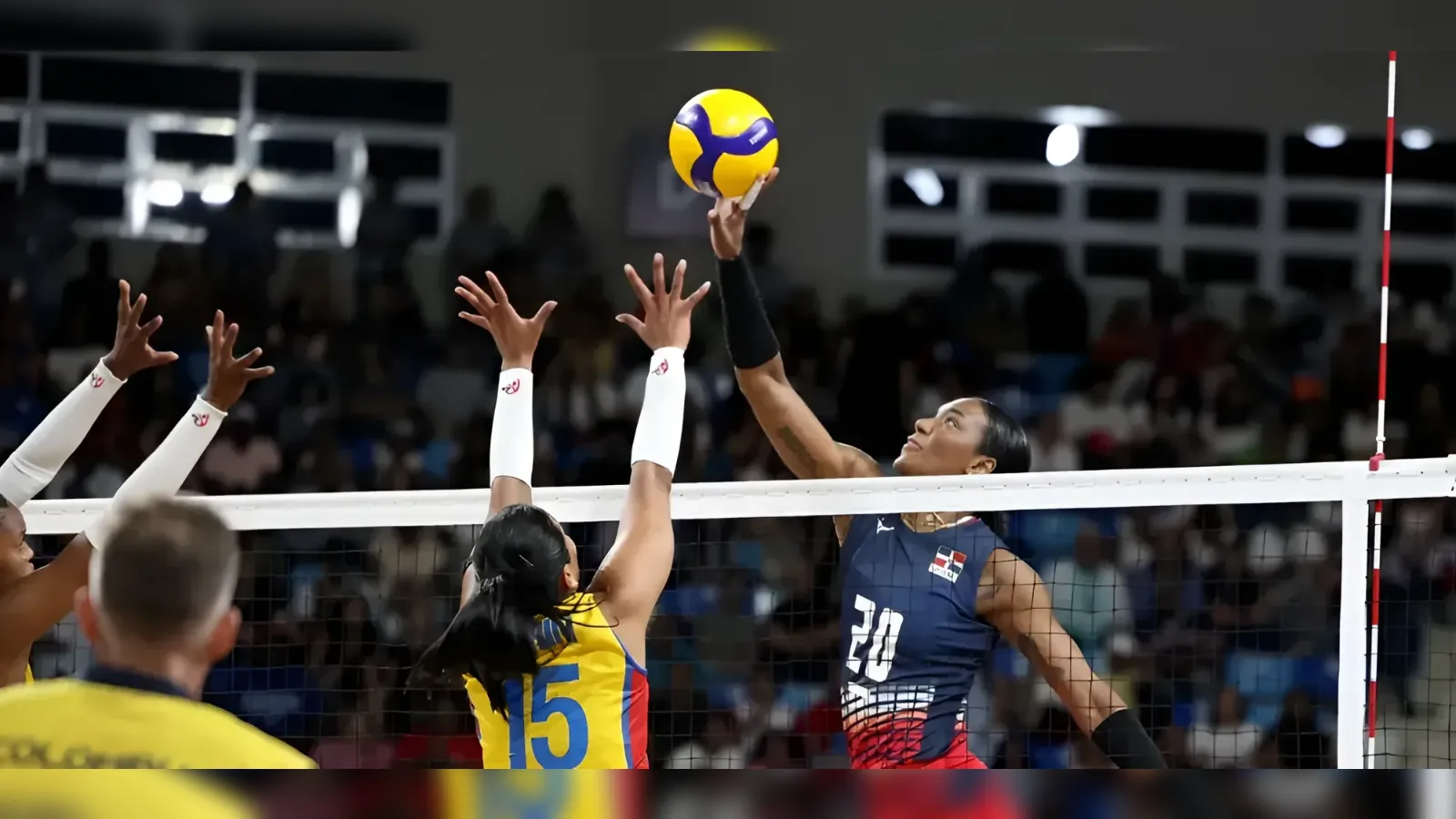 Volleyball action at the net: a player in navy (#30) spikes the ball while two blockers in yellow (#15) jump to block; crowd in the stands behind.