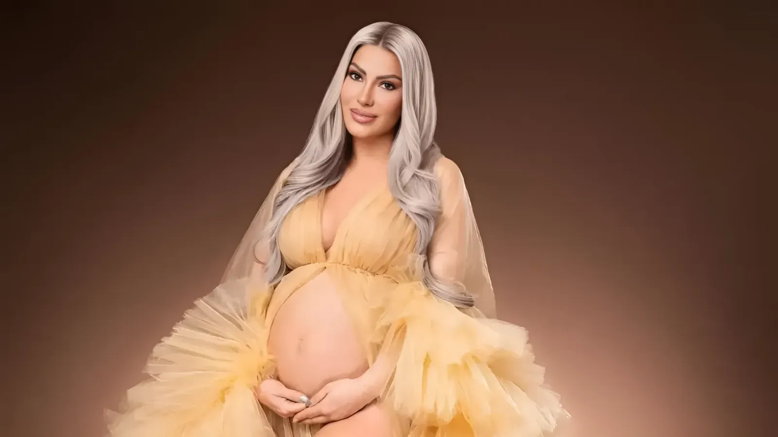 Pregnant woman in a flowing pale-yellow gown cradling her belly, with long platinum hair against a warm brown background.