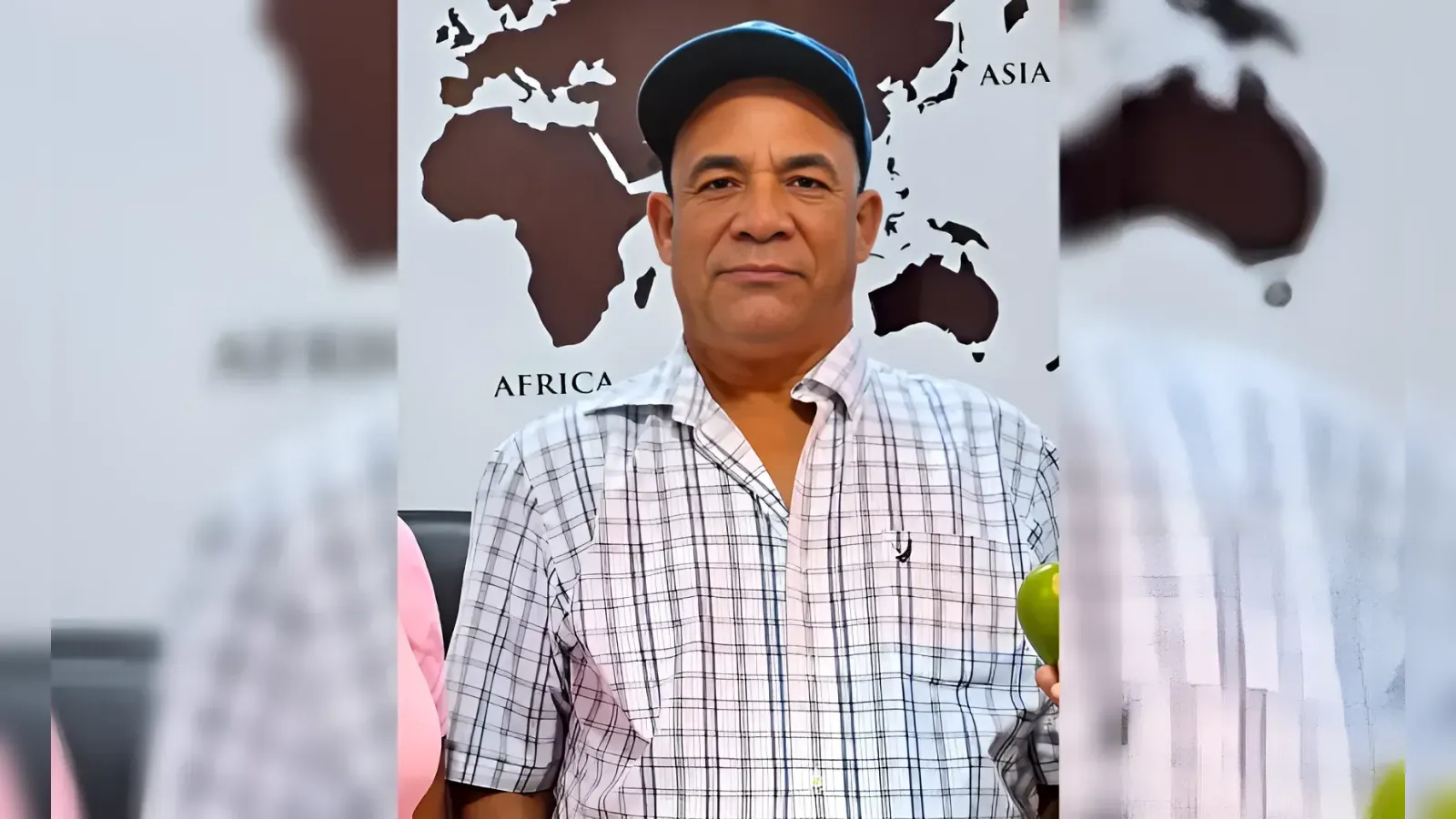 Man in a light plaid shirt and blue cap stands indoors with a world map backdrop showing Africa and Asia, holding a green apple in one hand.