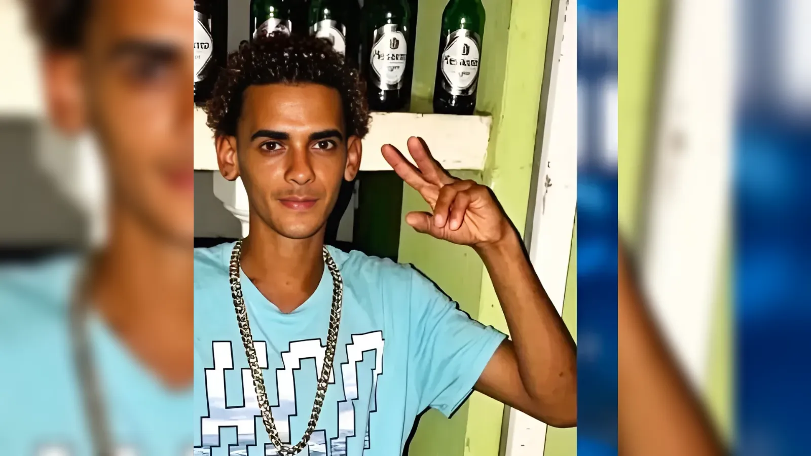 Young man with curly hair in a blue graphic T‑shirt and gold chains, flashing a peace sign beside bottles on a shelf behind him.