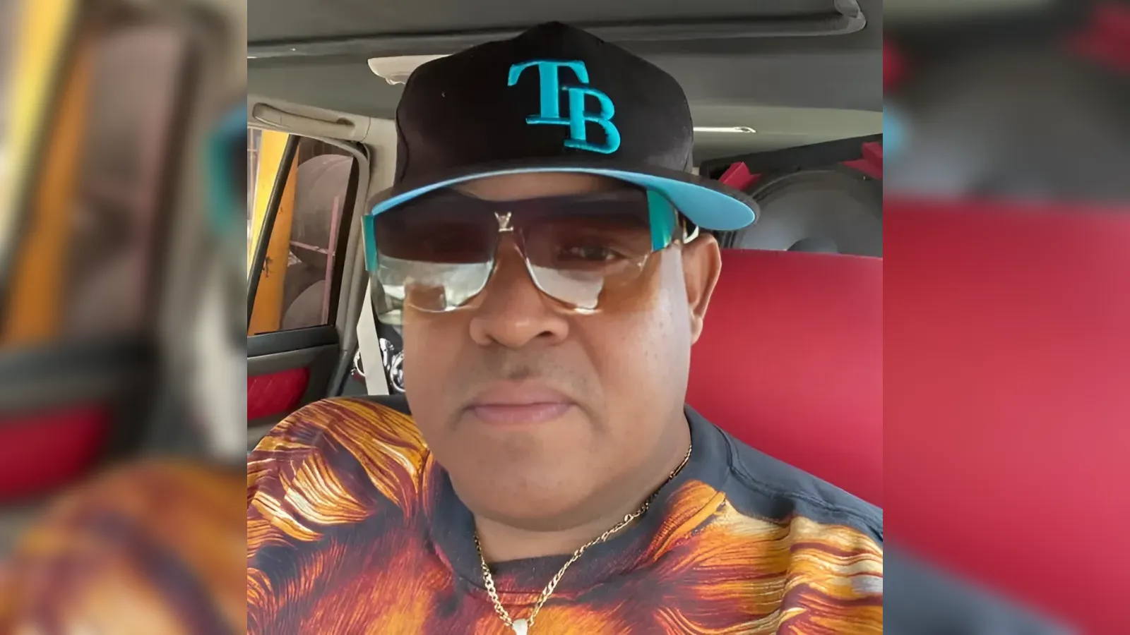 Close-up of a man in a car wearing a black cap with teal TB logo, oversized teal-accented sunglasses, and a colorful patterned shirt with a gold chain visible.