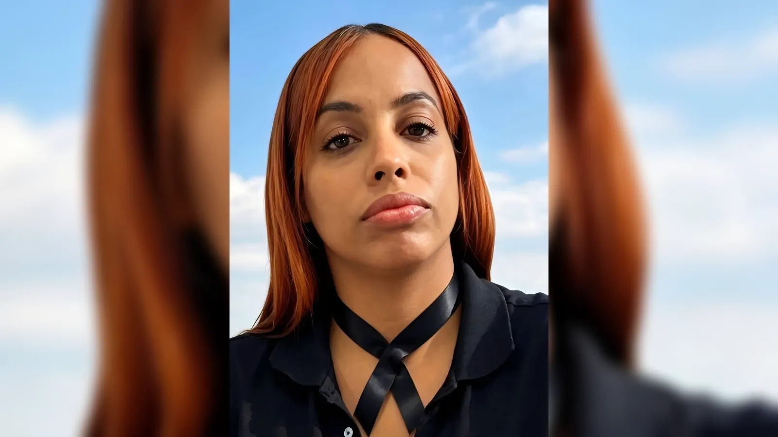 Close-up of a woman with orange hair outdoors, wearing a black crisscross neck scarf and dark shirt against a blue sky