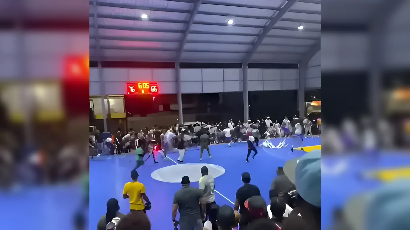 Crowd watches a sporting match on a bright blue indoor mat with a red glowing scoreboard overhead.