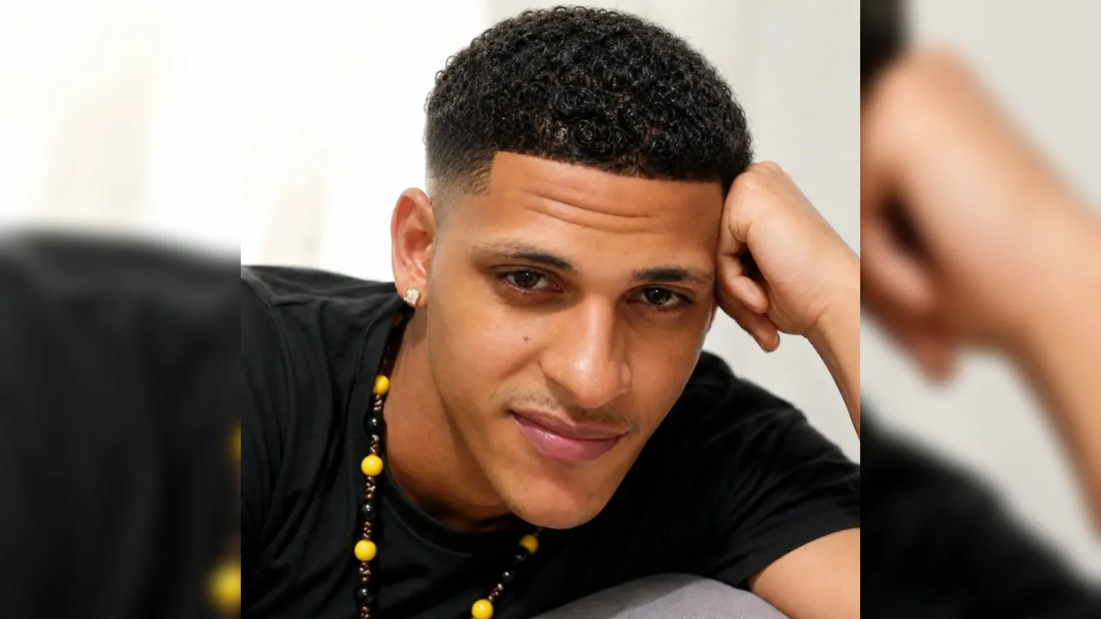 Close-up portrait of a man with short curly hair resting his head on his hand, wearing a black shirt and a yellow-black beaded necklace with an earring in his left ear.