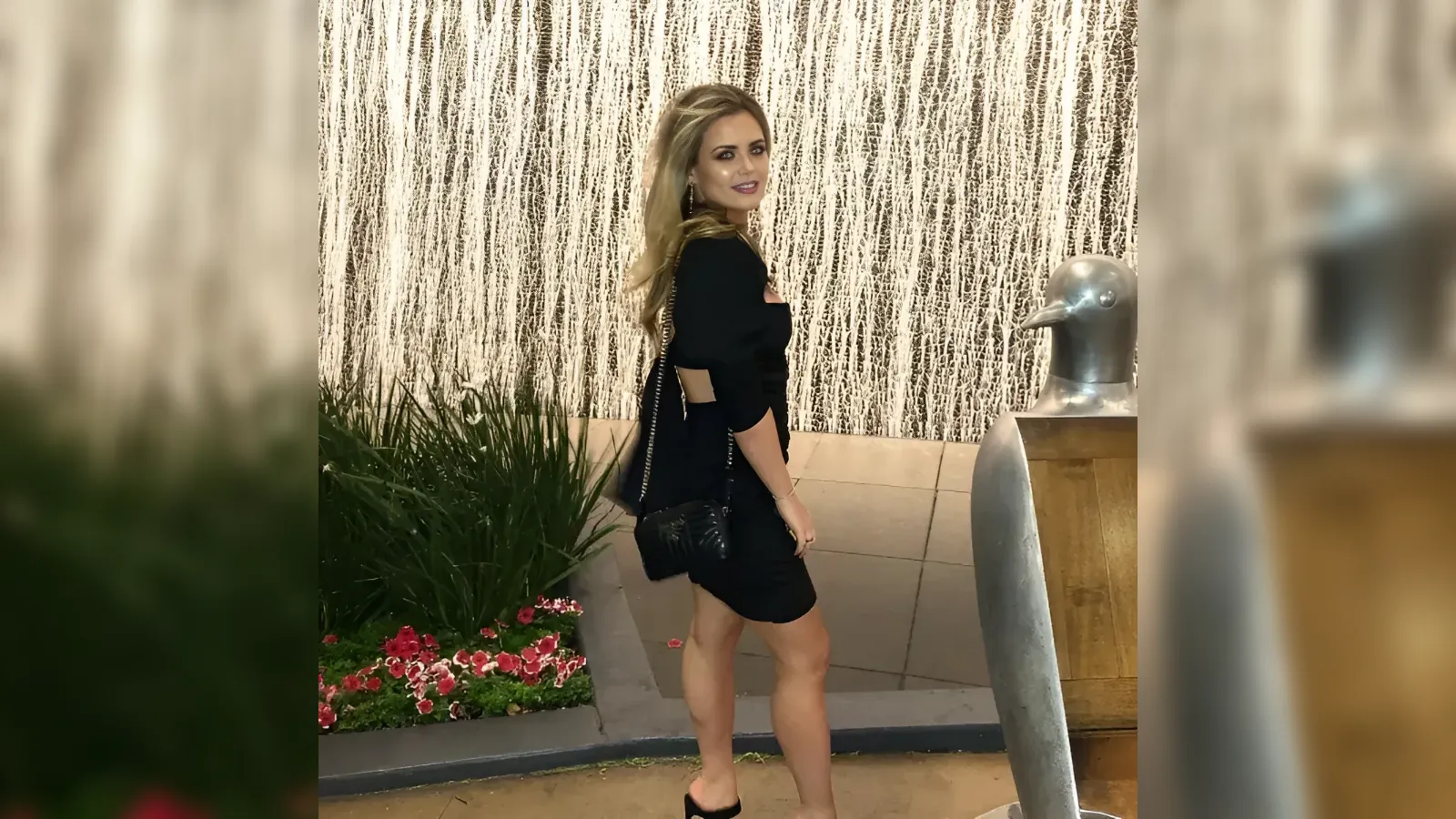 Woman in a black dress posing sideways beside a metallic duck sculpture outdoors, with a textured wall and greenery nearby.
