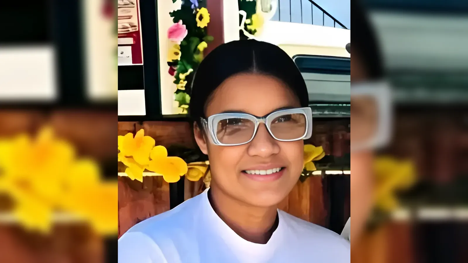 Person smiling at the camera, wearing white clothing and large white-framed glasses, with yellow flowers in the background.
