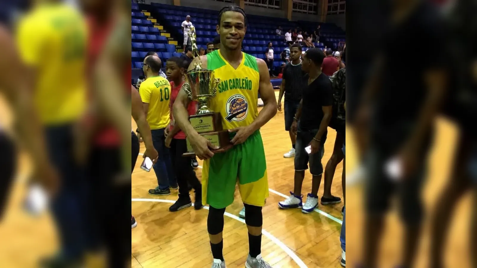 Basketball player in yellow-green San Carleño uniform holding a large trophy on a court, smiling after winning a championship.