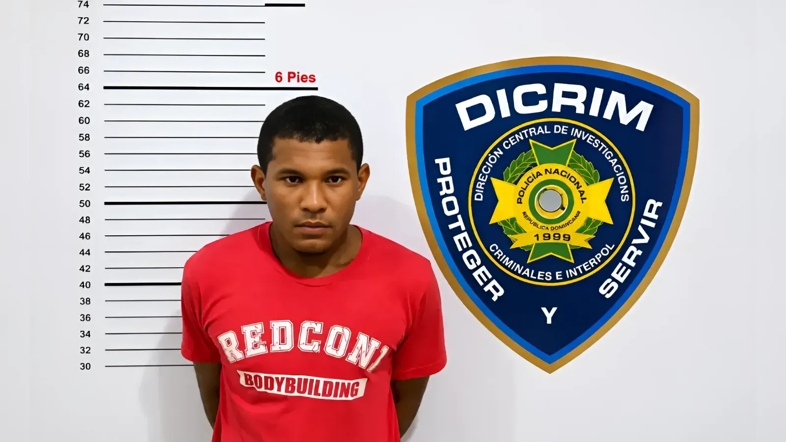 Mugshot-style photo of a man in a red t-shirt standing against a height chart, with a large police emblem on the wall to the right.