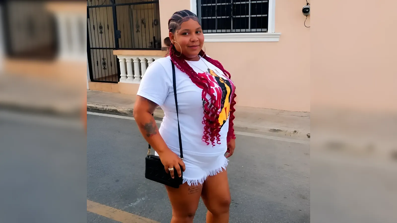 Woman standing on a city street in a white T‑shirt and white frayed shorts, with red braids and hoop earrings.