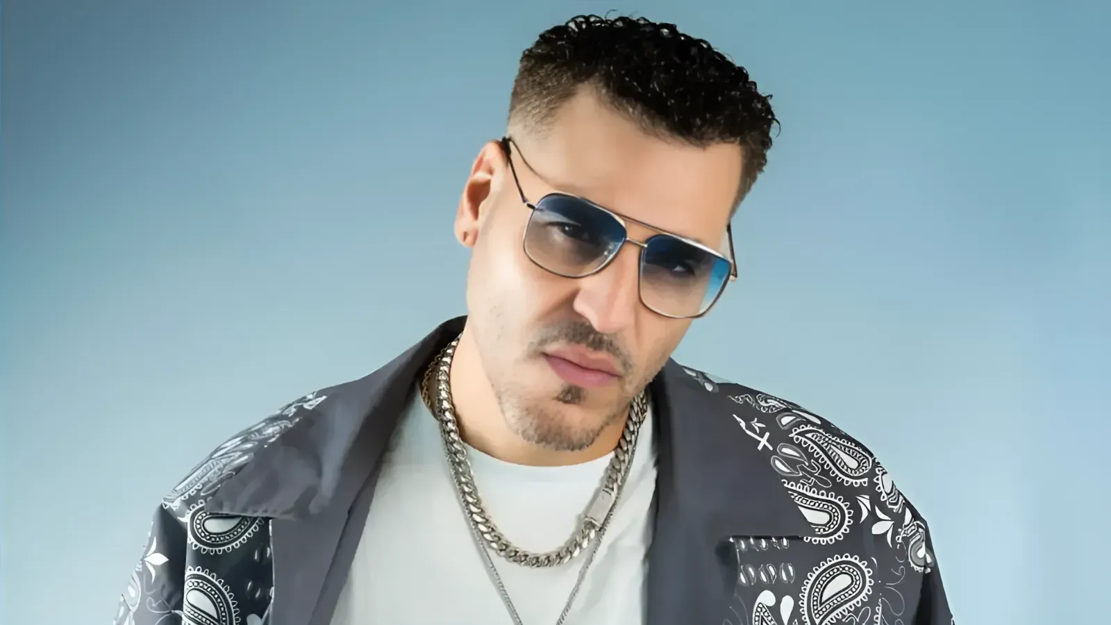 Portrait of a man wearing aviator sunglasses, a patterned dark shirt, and silver chains, looking at the camera.