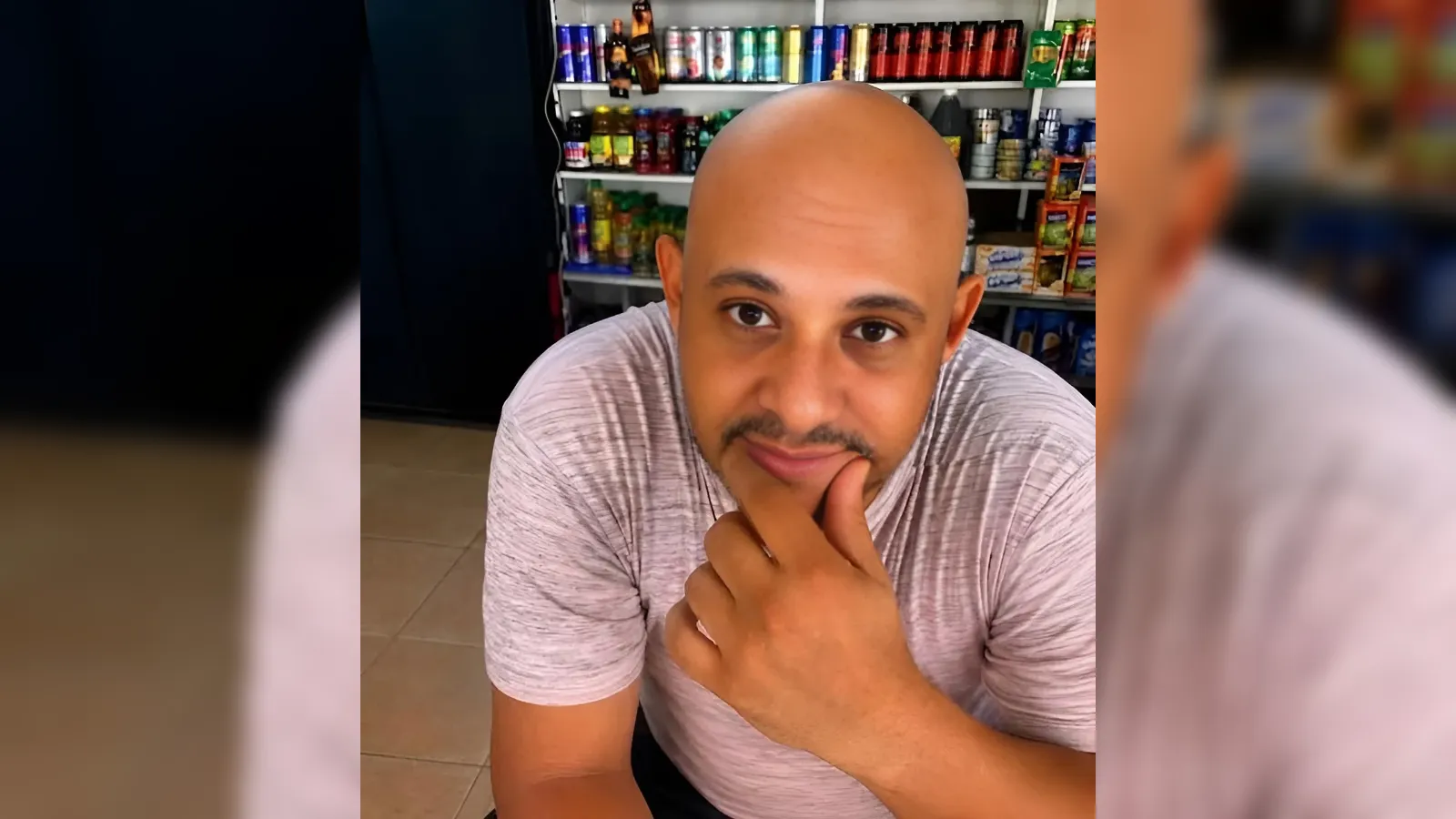 Man with a shaved head in a store, resting his chin on his hand and smiling at the camera.