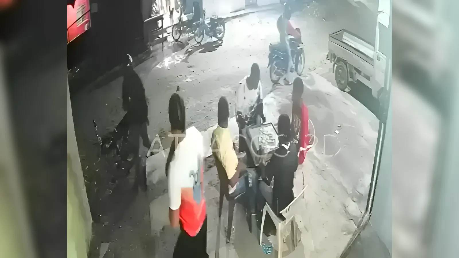 Nighttime street scene with people gathered around a small seating area outside a shop; bicycles and a truck are nearby.