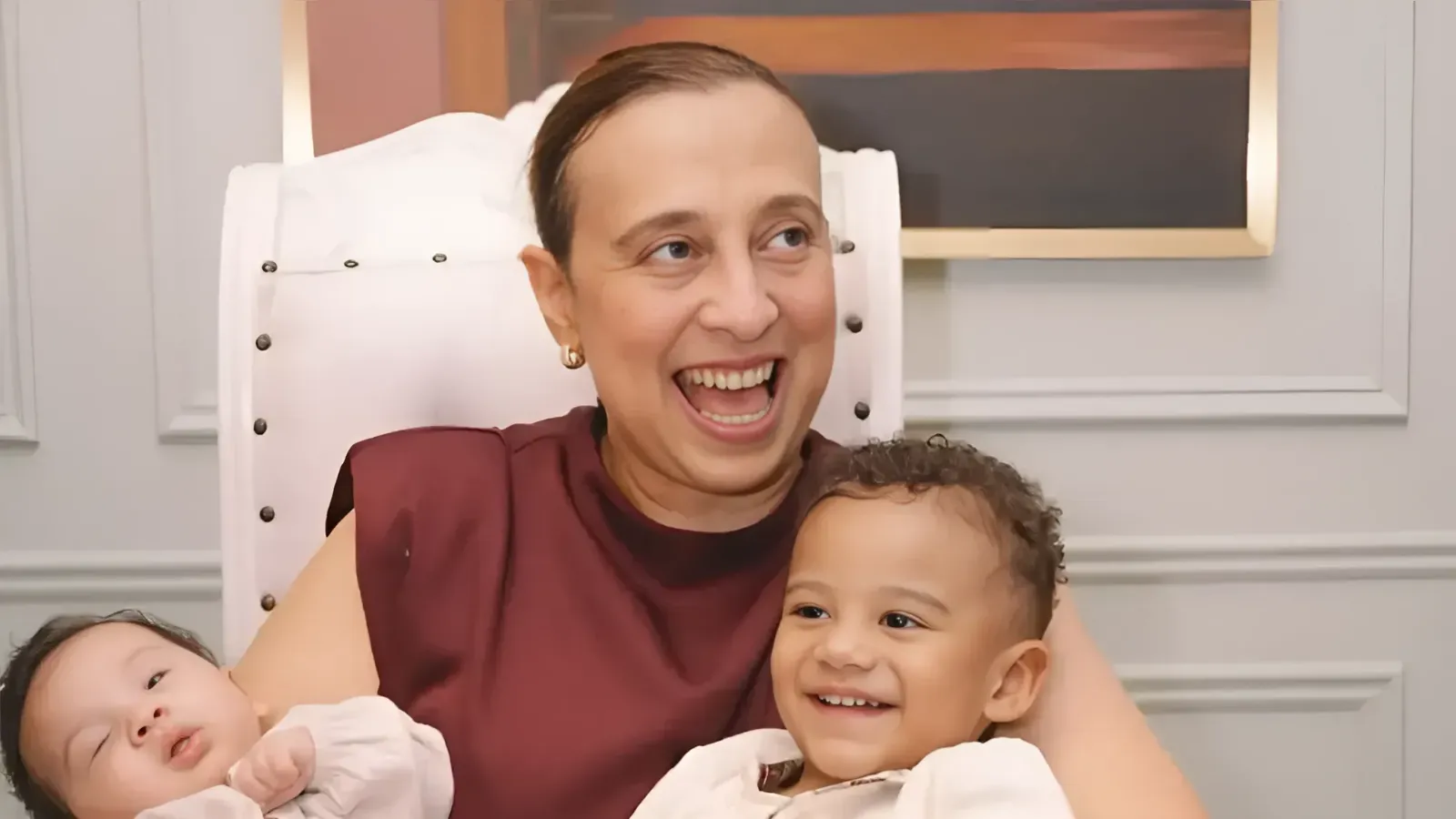 Woman smiling and laughing while holding two babies, one asleep on her left and one awake on her right, in a warmly lit room.