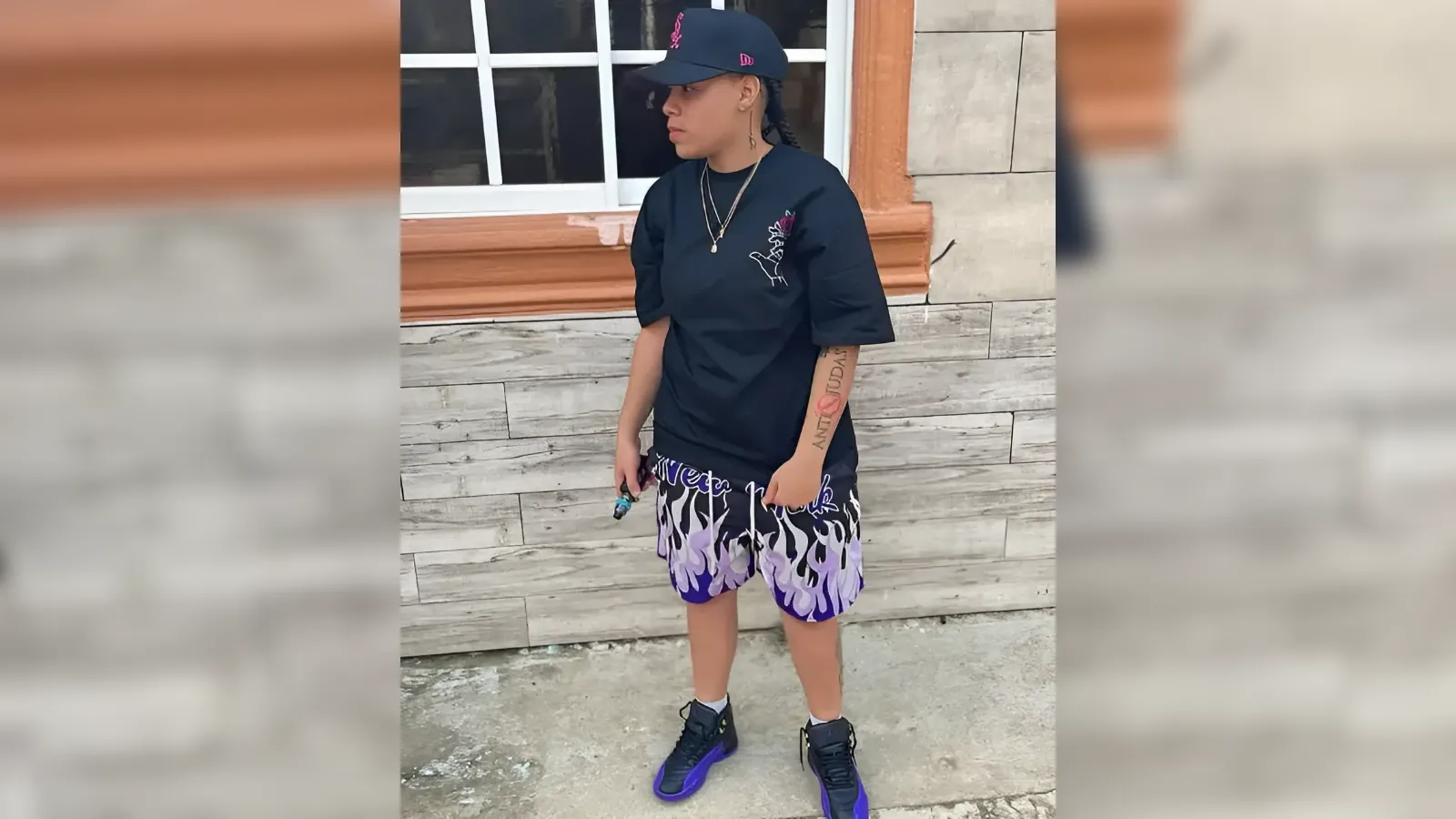 Person standing in front of a stone wall wearing a black oversized T-shirt, purple flame shorts, black cap, sneakers, and gold necklaces; visible tattoo on left forearm.
