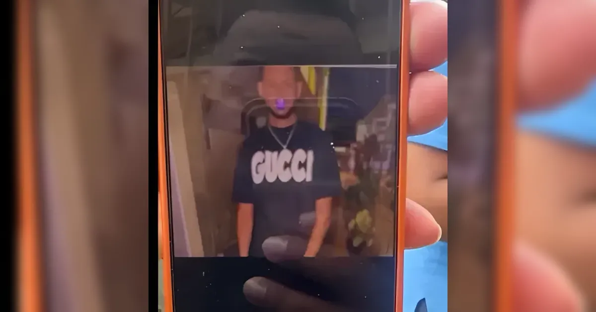 Person wearing a black Gucci t‑shirt is shown on a smartphone screen being held up, with a blurred indoor background behind him.