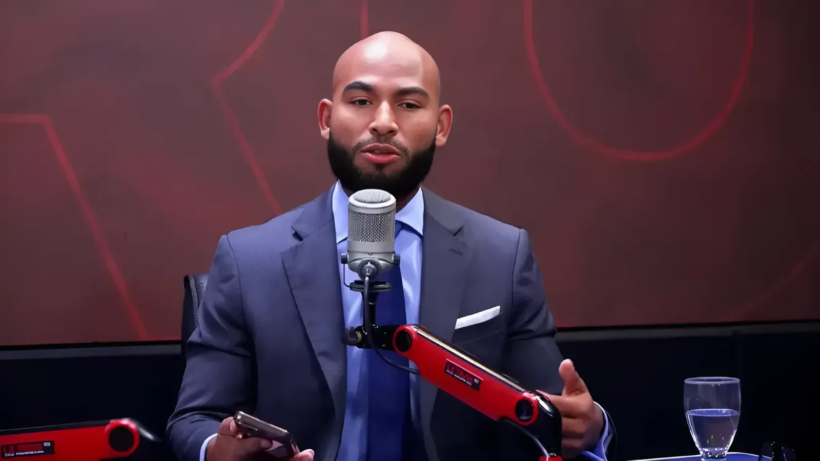 Professional podcast recording scene: man in a blue suit speaks into a large studio microphone, with a red boom arm nearby and a glass of water on the desk