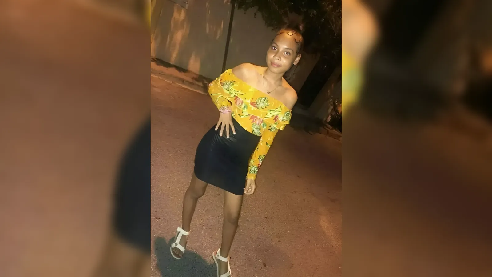 A young woman posing on a street at night wearing a yellow off-shoulder floral top and a black mini skirt with white sandals.
