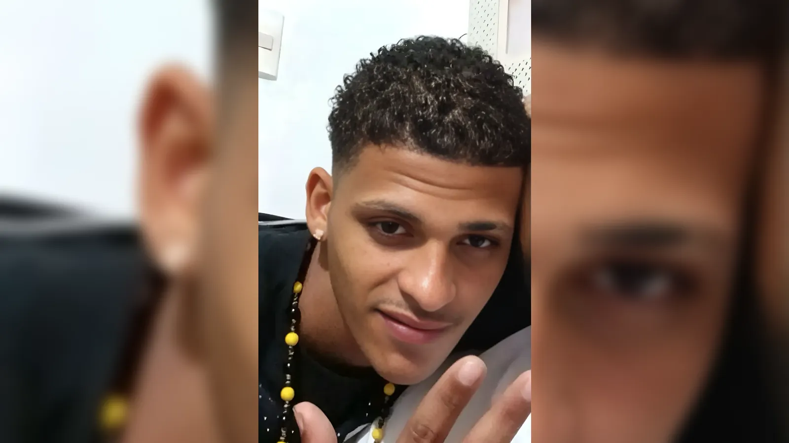 Selfie of a young man with short curly hair, looking into the camera and showing two fingers in a peace sign, wearing a beaded necklace.