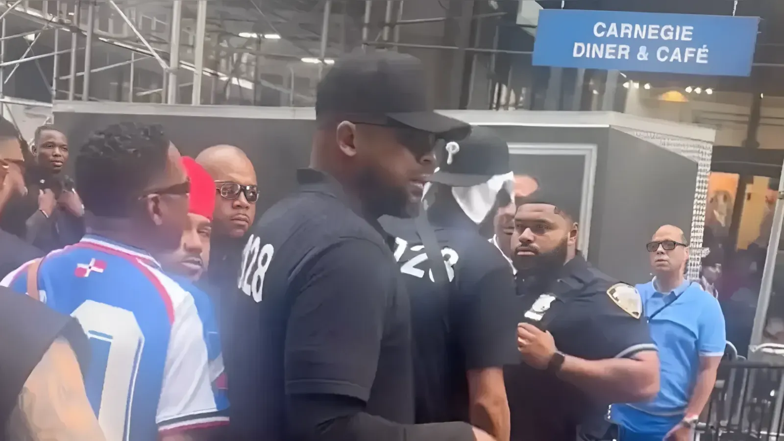 Man in a black cap and black shirt (number 28 on back) stands among a crowd outside Carnegie Diner & Café, with a police officer nearby.