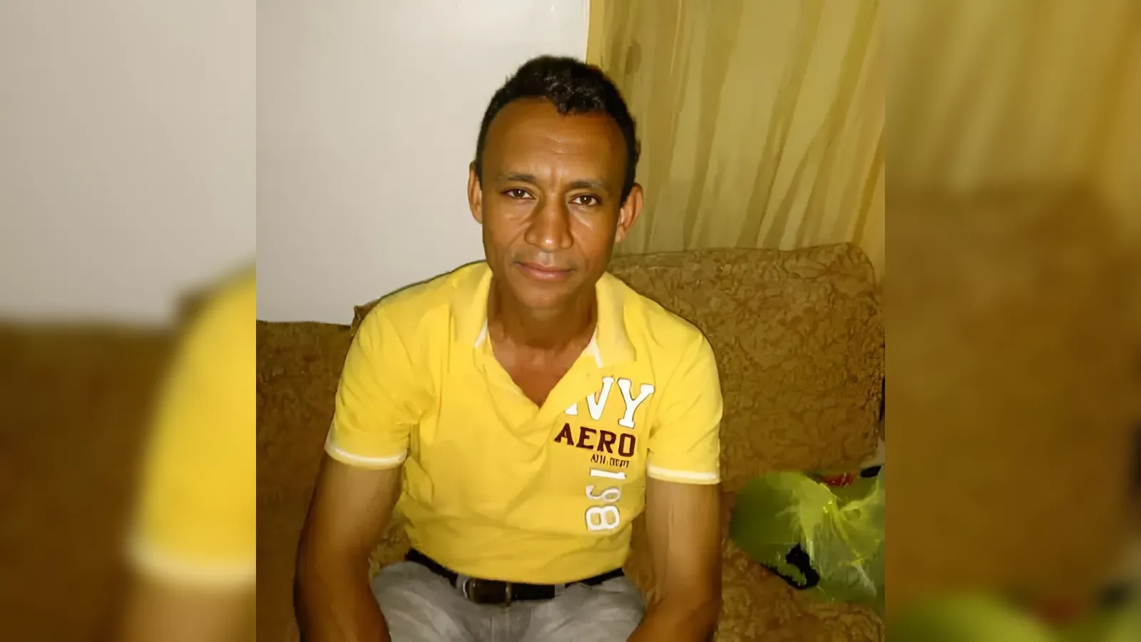 Man with short dark hair in a yellow polo shirt sitting on a patterned sofa in a living room, looking at the camera.