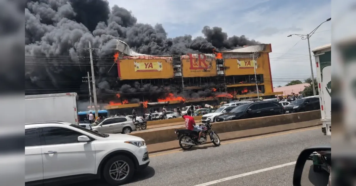 Commercial building on a highway is burning with huge flames and black smoke, cars and motorcycles stuck in traffic.
