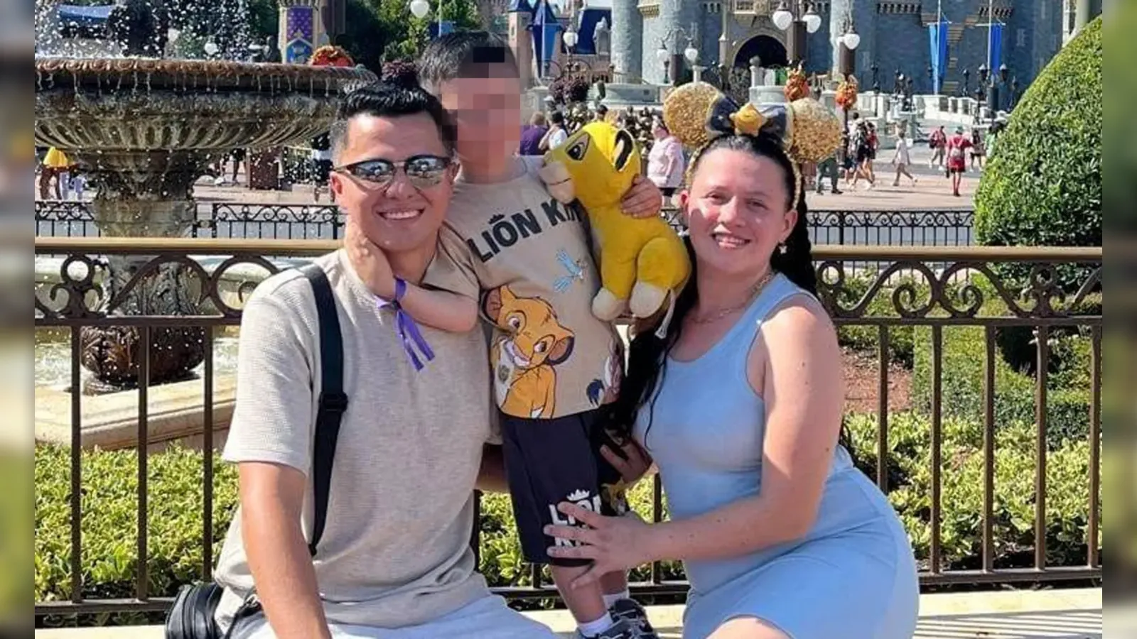 Three people pose by a theme park fountain: a man in a gray shirt holds a child with a Simba plush, while a woman in a blue dress wears Minnie ears and smiles.