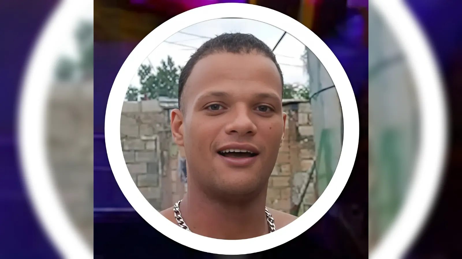 Close-up portrait of a smiling man wearing a silver chain necklace, outdoors behind a brick wall, inside a white circular frame.