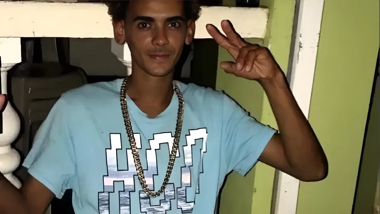 Young man in a light blue graphic T‑shirt and gold chain posing with peace signs indoors near a stair railing.
