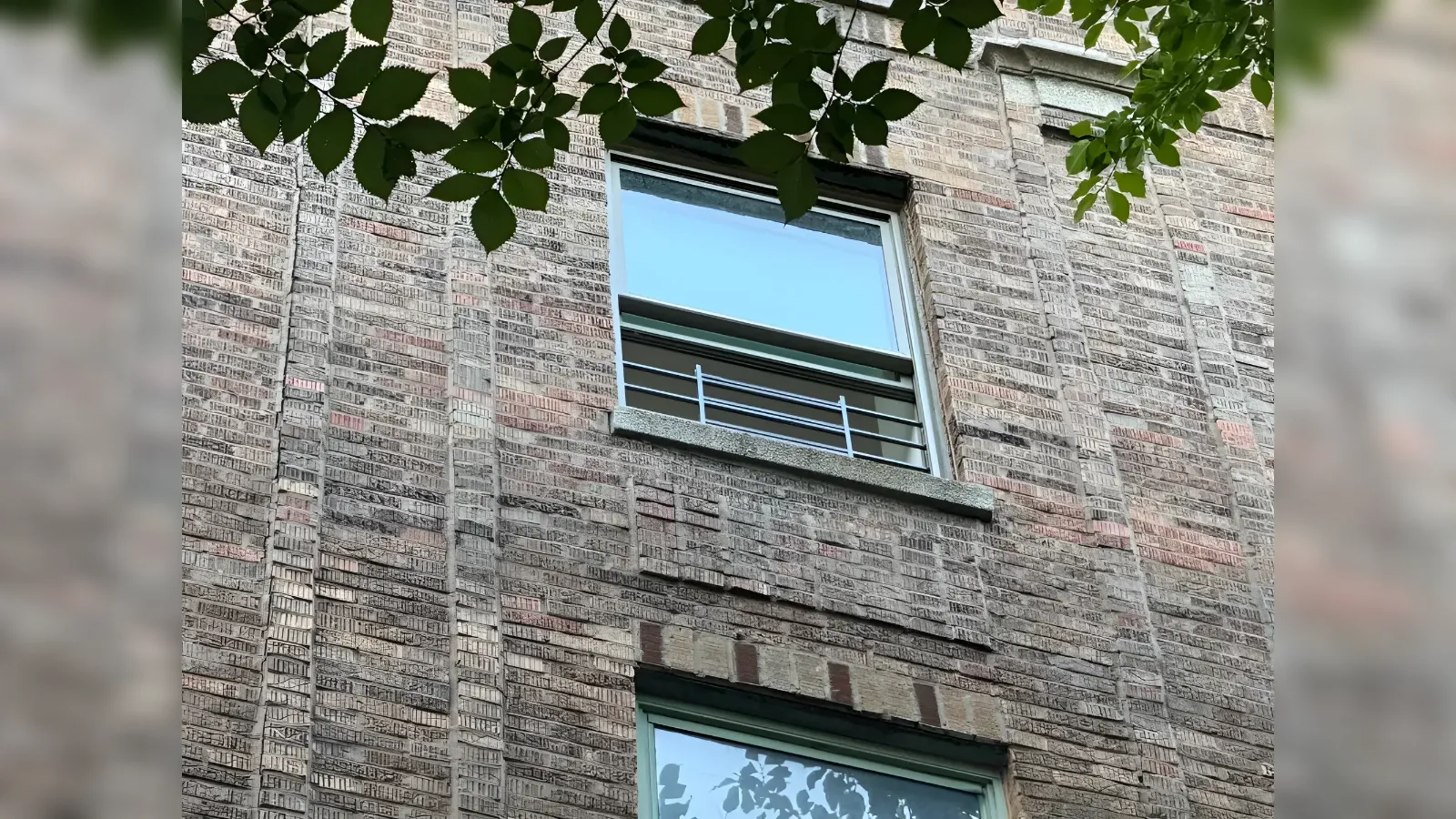 Brick building window with a gray stone sill and slim horizontal railing, framed by overhanging green leaves.
