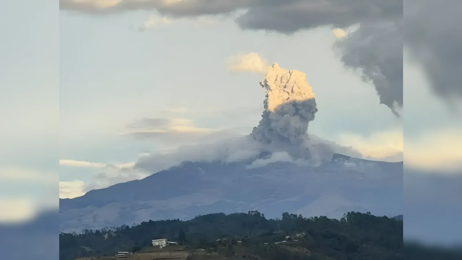 Volcanic eruption plume rising from a mountain, ash and smoke above a hilly landscape with trees and scattered buildings below.