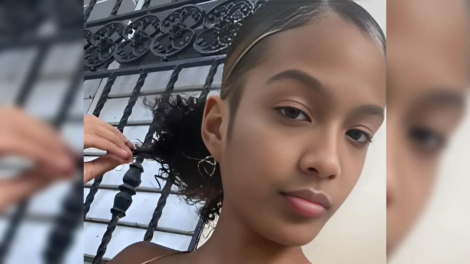 Close-up of a young woman posing for a selfie while a hand styles her curly hair beside a decorative wrought-iron gate.