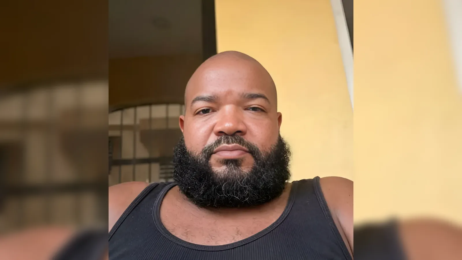 Close-up of a bald man with a thick beard wearing a black tank top, looking at the camera indoors with a yellow wall behind him.