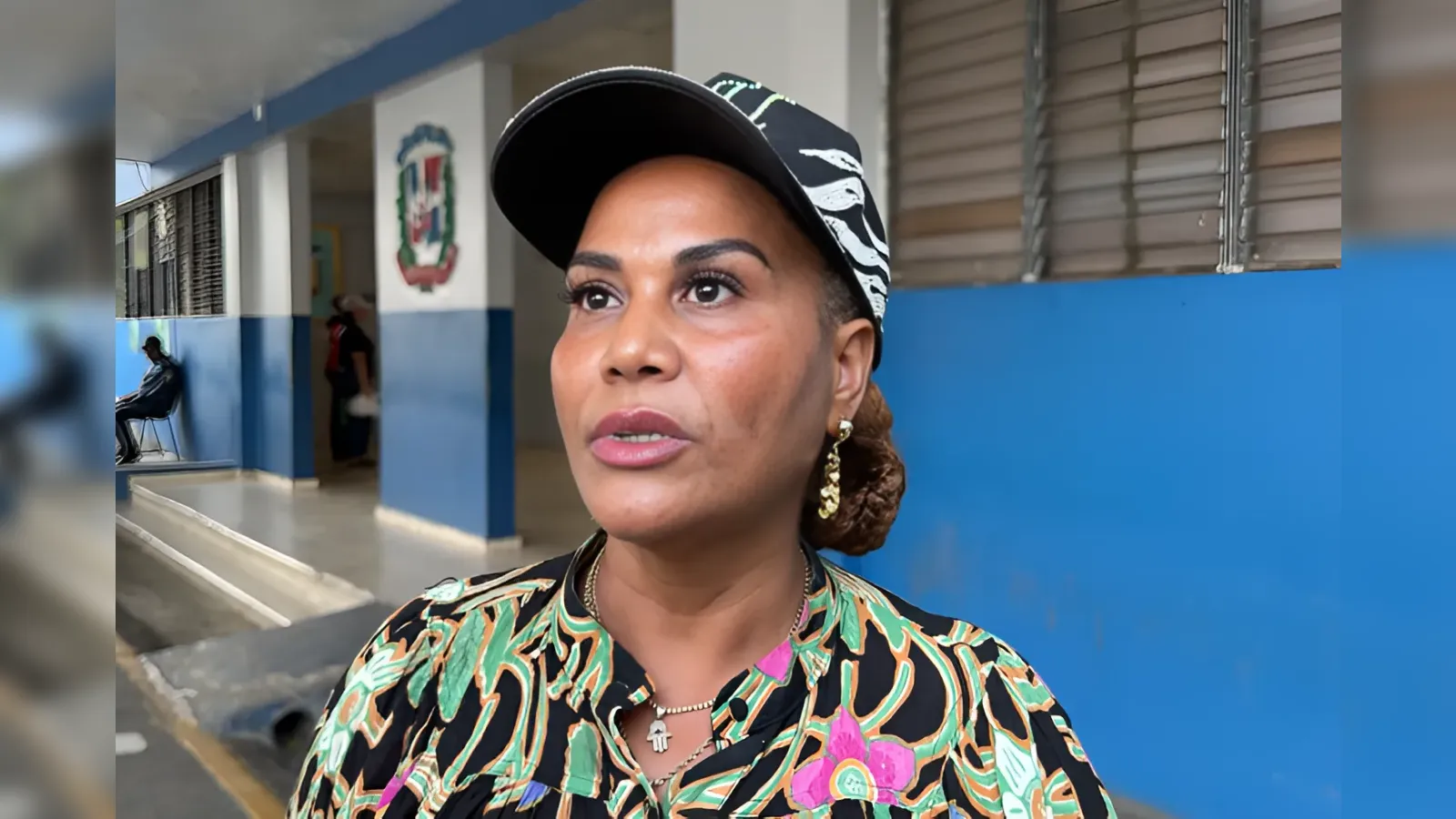 Close-up of a woman wearing a colorful tropical shirt and a black cap, speaking in an indoor hallway with blue walls behind her.