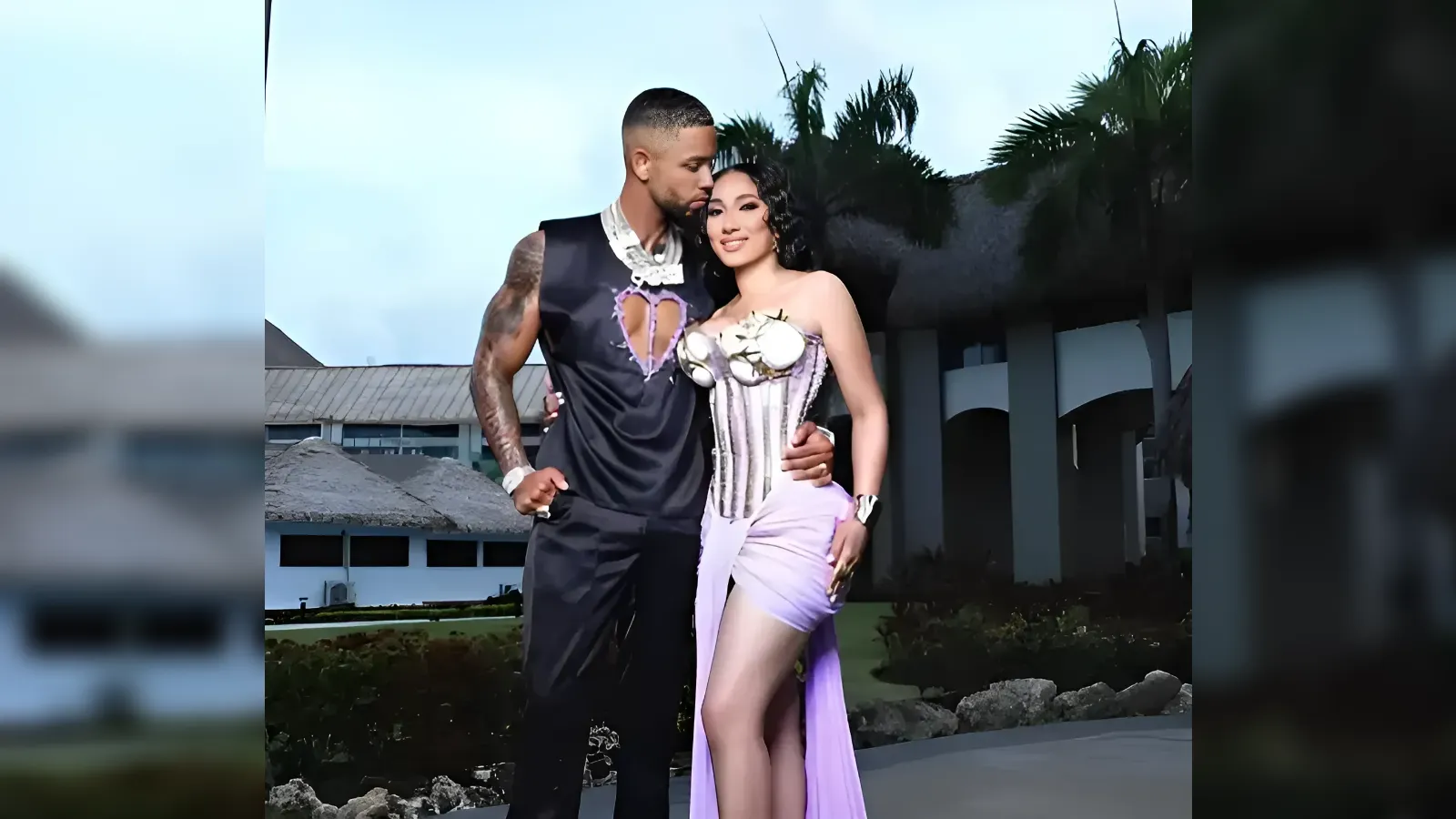 Couple posing together outdoors in fashionable outfits; man with tattoos in a sleeveless black top and the woman in a lavender corset-style dress, palm trees in the background