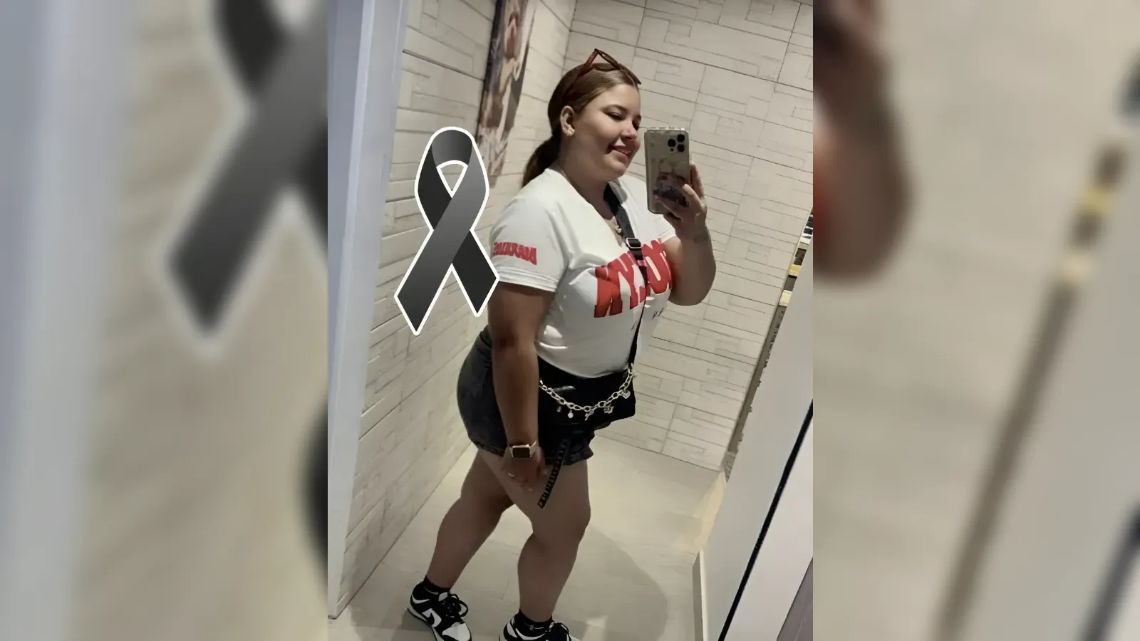 Person posing for a mirror selfie in a tiled room, wearing a white T‑shirt, black shorts with a chain belt, sneakers, with a large black awareness ribbon overlay.