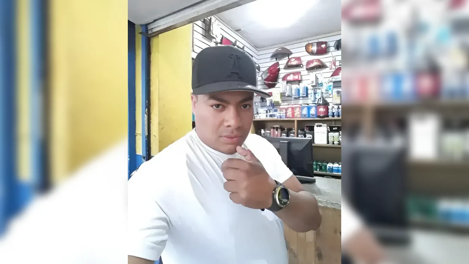 Man in a black cap and white shirt points at the camera inside an auto-parts shop (shelves of car parts in background).