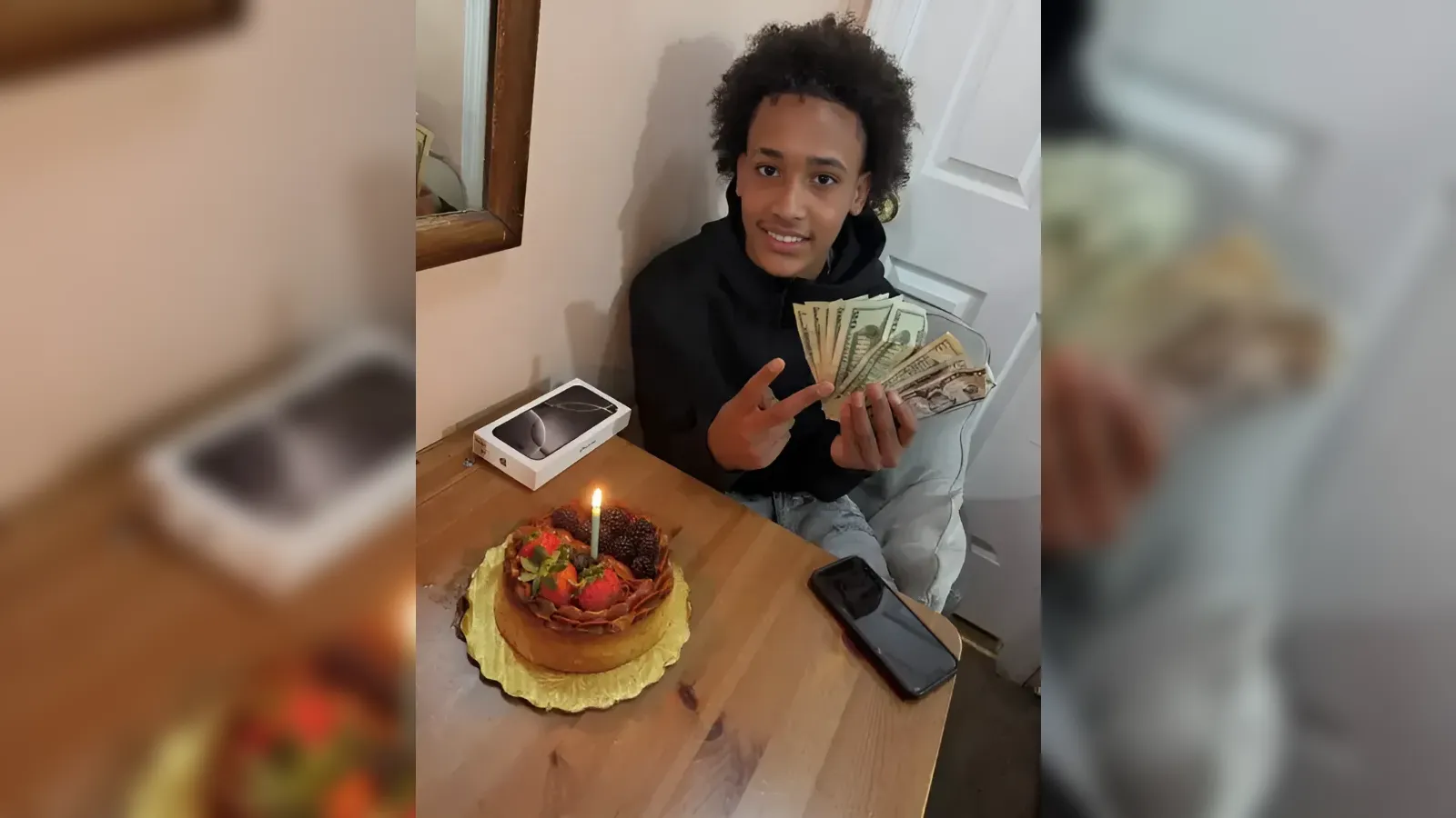 Young person in a black hoodie holds a fan of cash and smiles at the camera beside a birthday cake with a lit candle on a wooden table.
