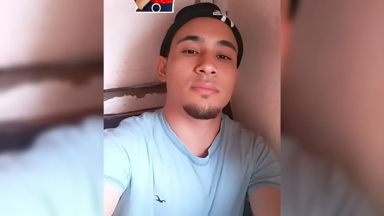Young man wearing a black cap backward and a light blue T-shirt, leaning against a pink wall and looking at the camera