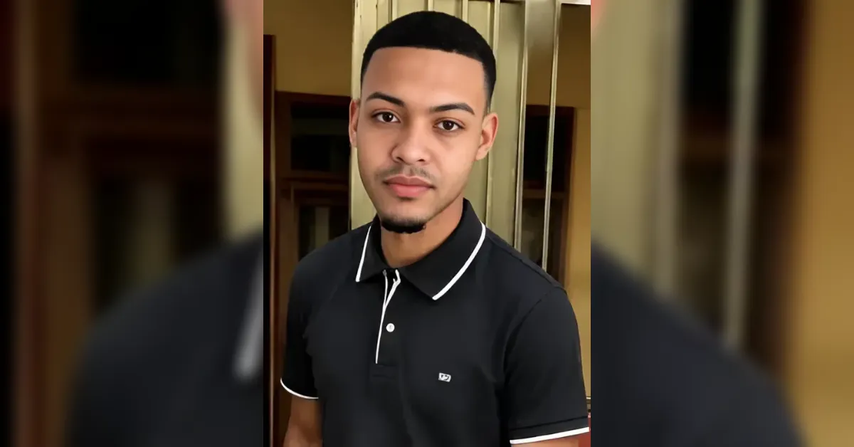 Portrait of a young man in a black polo shirt with white trim, standing indoors near a doorway.