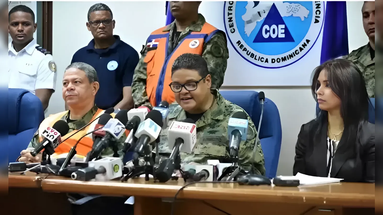 Military officials at a press conference with numerous microphones on the table, COE emblem in the background, man in uniform speaking while others listen.