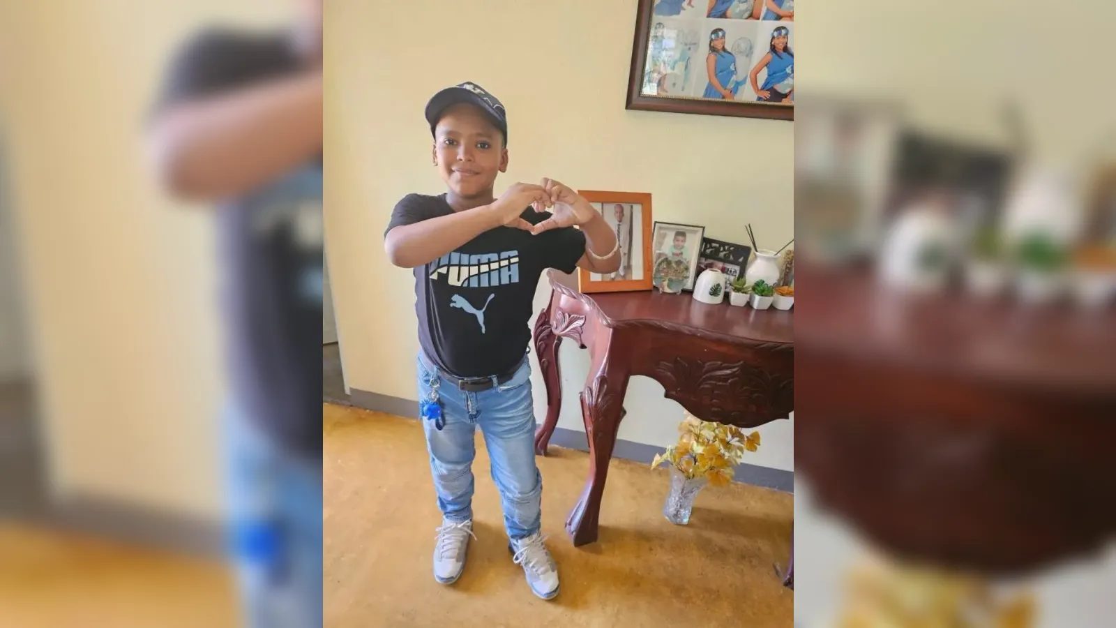 Boy wearing a cap and black t-shirt forms a heart shape with his hands indoors, smiling at the camera.
