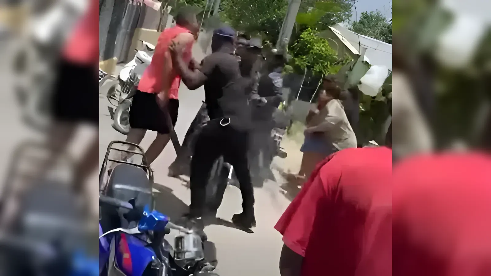 Outdoor street scene of a man in an orange shirt being restrained by a person in black while others watch; a scooter is visible on the left.