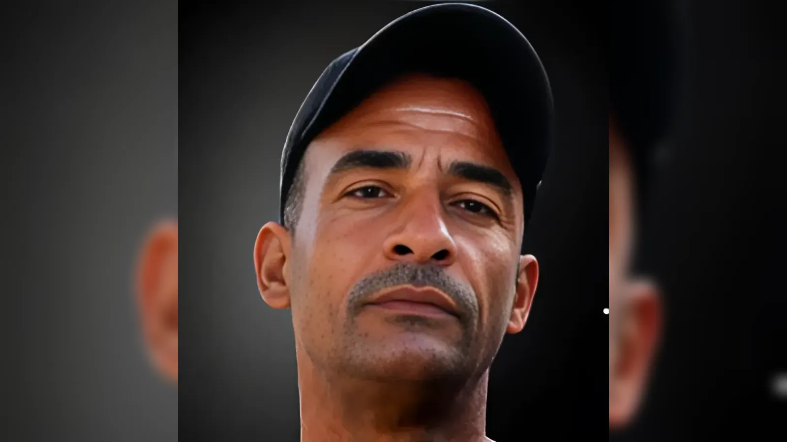 Close-up of a man wearing a black cap, looking straight ahead with a neutral expression against a dark background.
