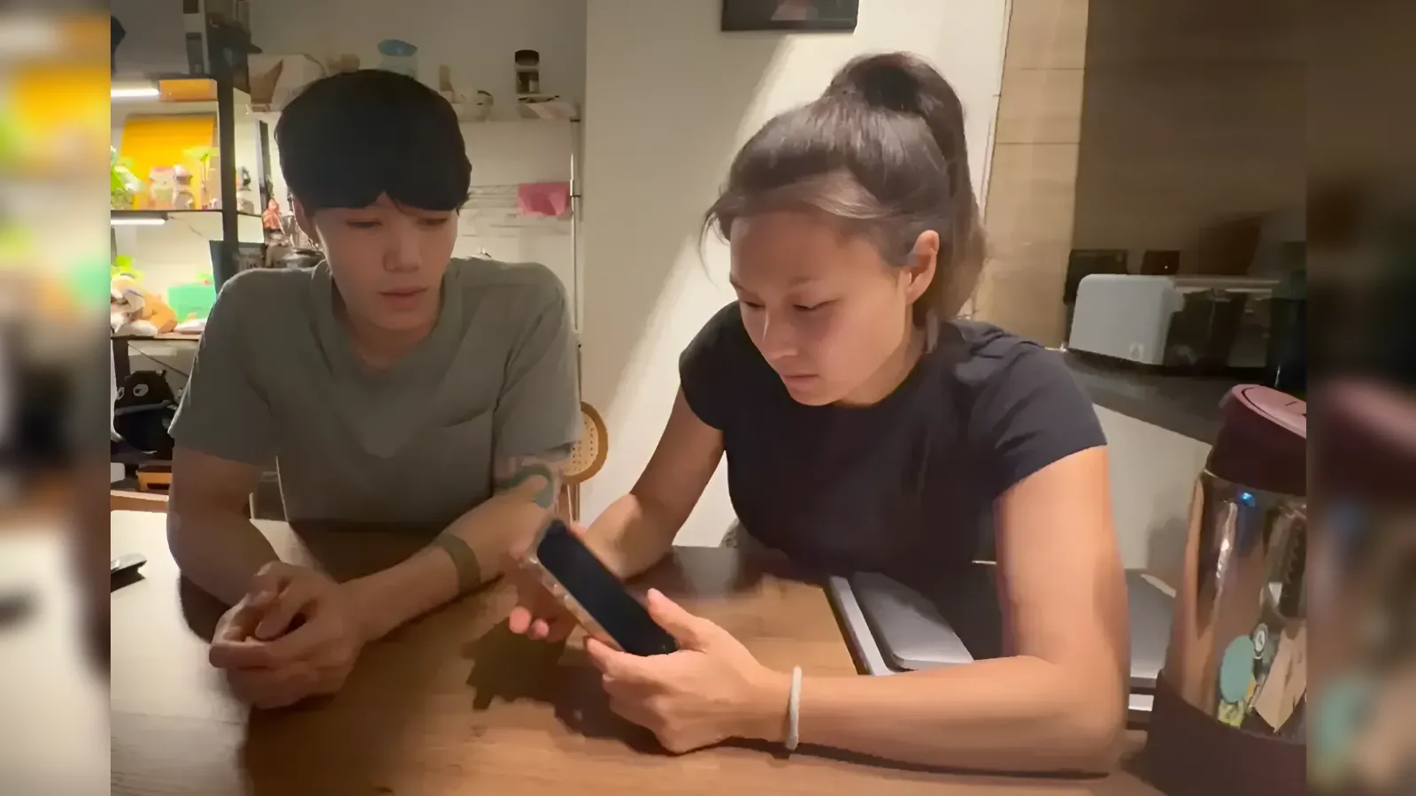 Two people sit at a wooden table, focused on a smartphone the woman is holding.