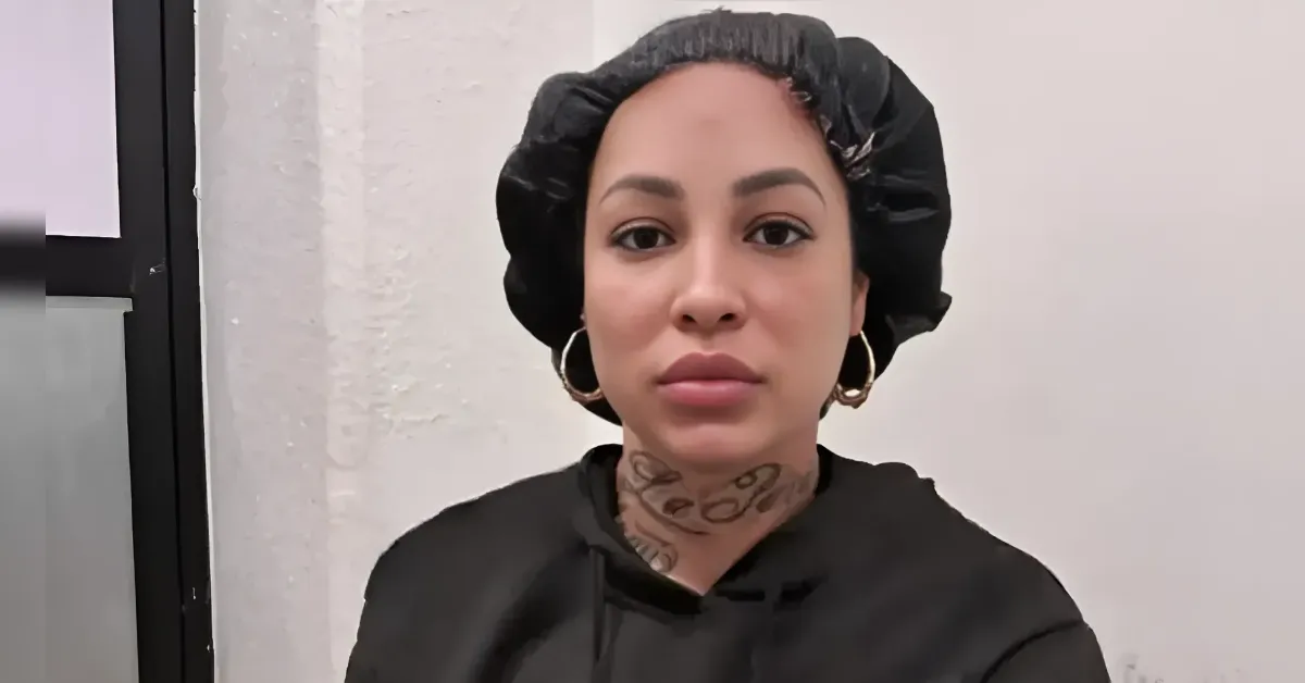Close-up of a woman wearing a black satin bonnet and hoodie, with gold hoop earrings and a neck tattoo, facing the camera.