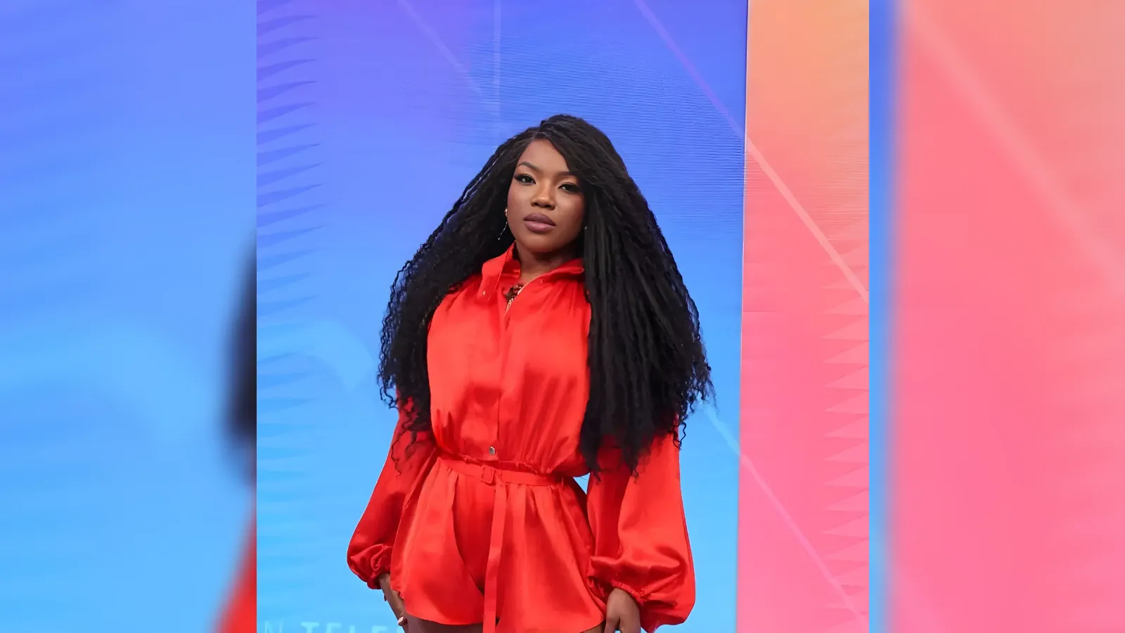 Woman with long braided black hair wearing a bright red satin romper stands against a blue to pink gradient backdrop on a stage.
