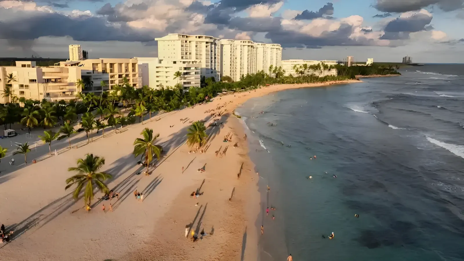 Aerial view of a tropical beach resort: white hotels along the shore, palm trees, and people on the sandy beach by the turquoise sea.
