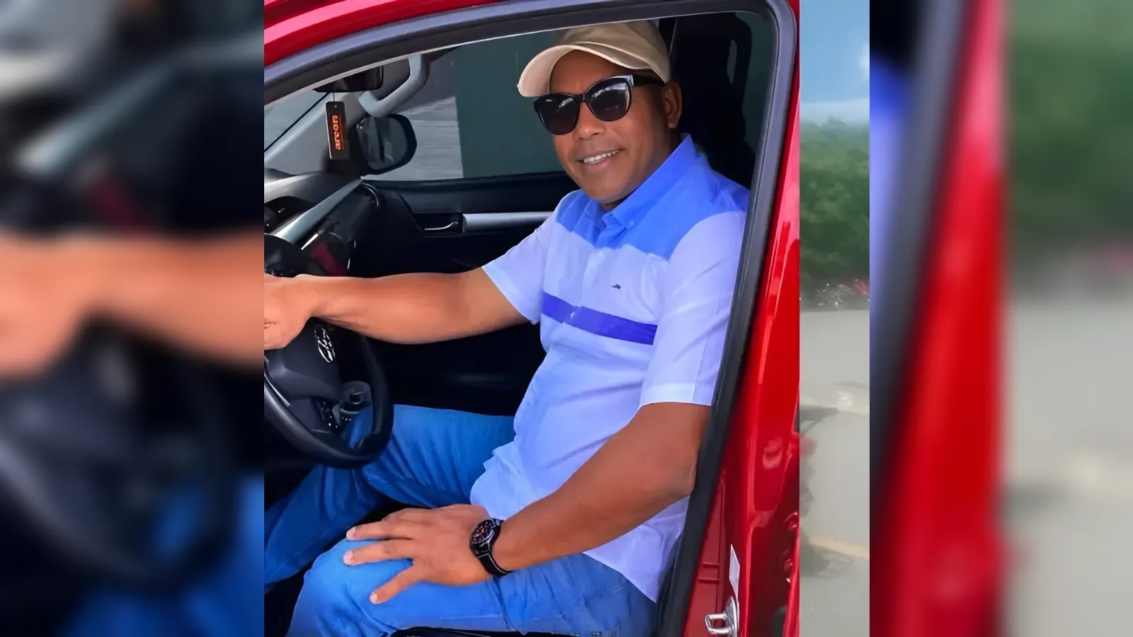 Man wearing cap and sunglasses sits in a red car, hands on the steering wheel, smiling at the camera (driver photo).